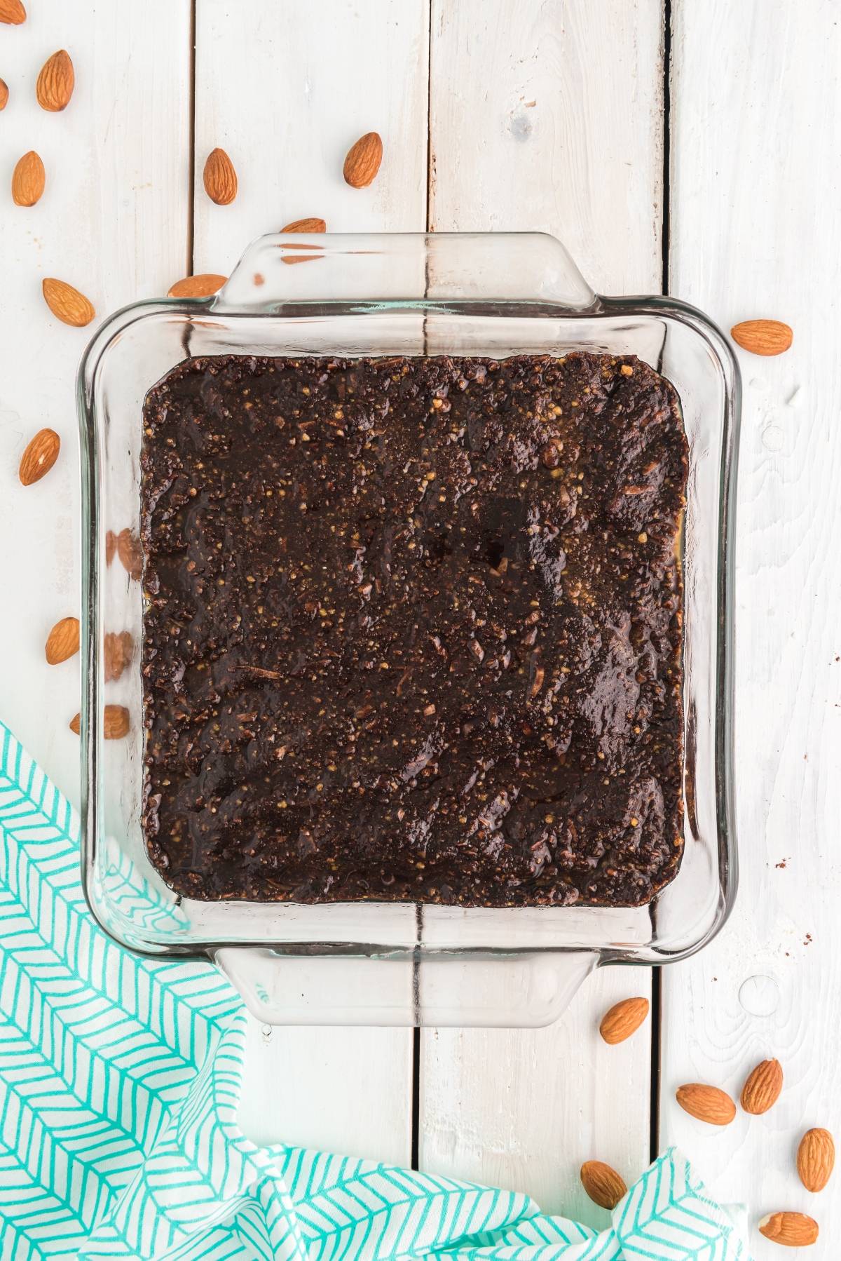 A glass dish with chocolate brownies, reminiscent of a classic Canadian dessert, sits on a white wooden surface with scattered almonds and a green patterned cloth.