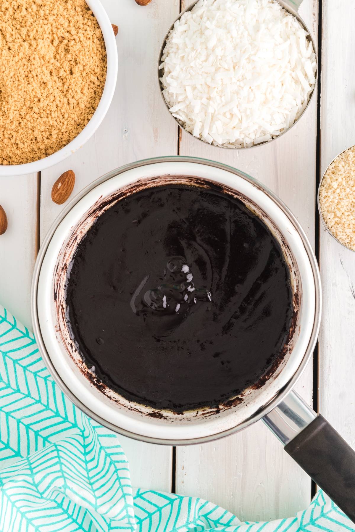 Saucepan with melted chocolate, surrounded by bowls of coconut flakes, graham crumbs, and nuts on a white table—perfect ingredients for making classic Canadian dessert Nanaimo bars, a delicious no bake dessert.