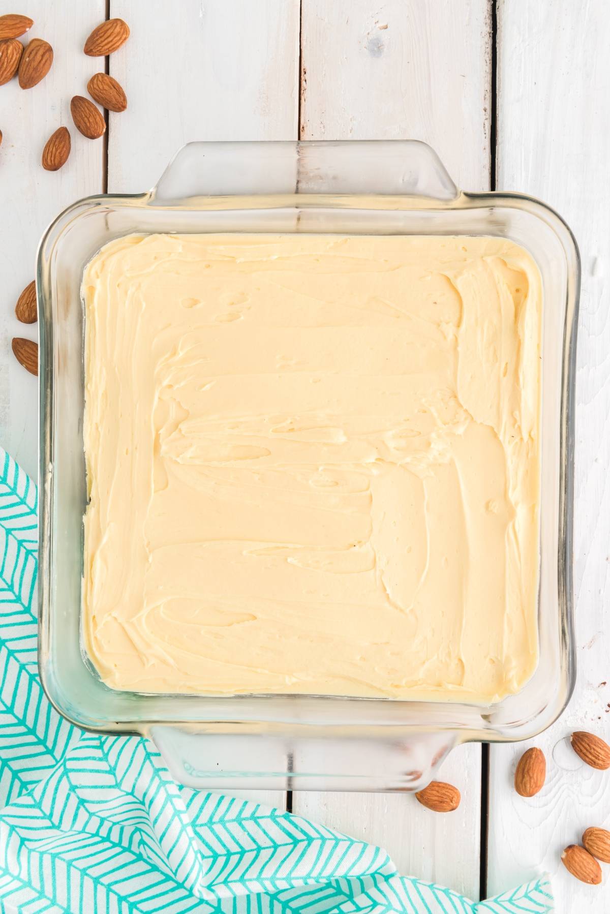 A glass baking dish with smooth, creamy batter—perfect for a layered bar recipe—surrounded by almonds on a white wooden surface.