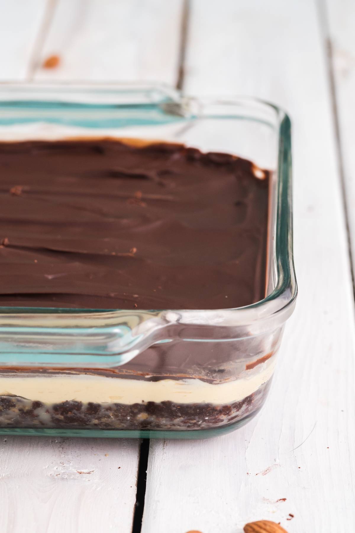 A glass dish with layered dessert featuring a chocolate top, creamy middle, and crumbly base—these Nanaimo bars are a classic Canadian dessert that freezes well.