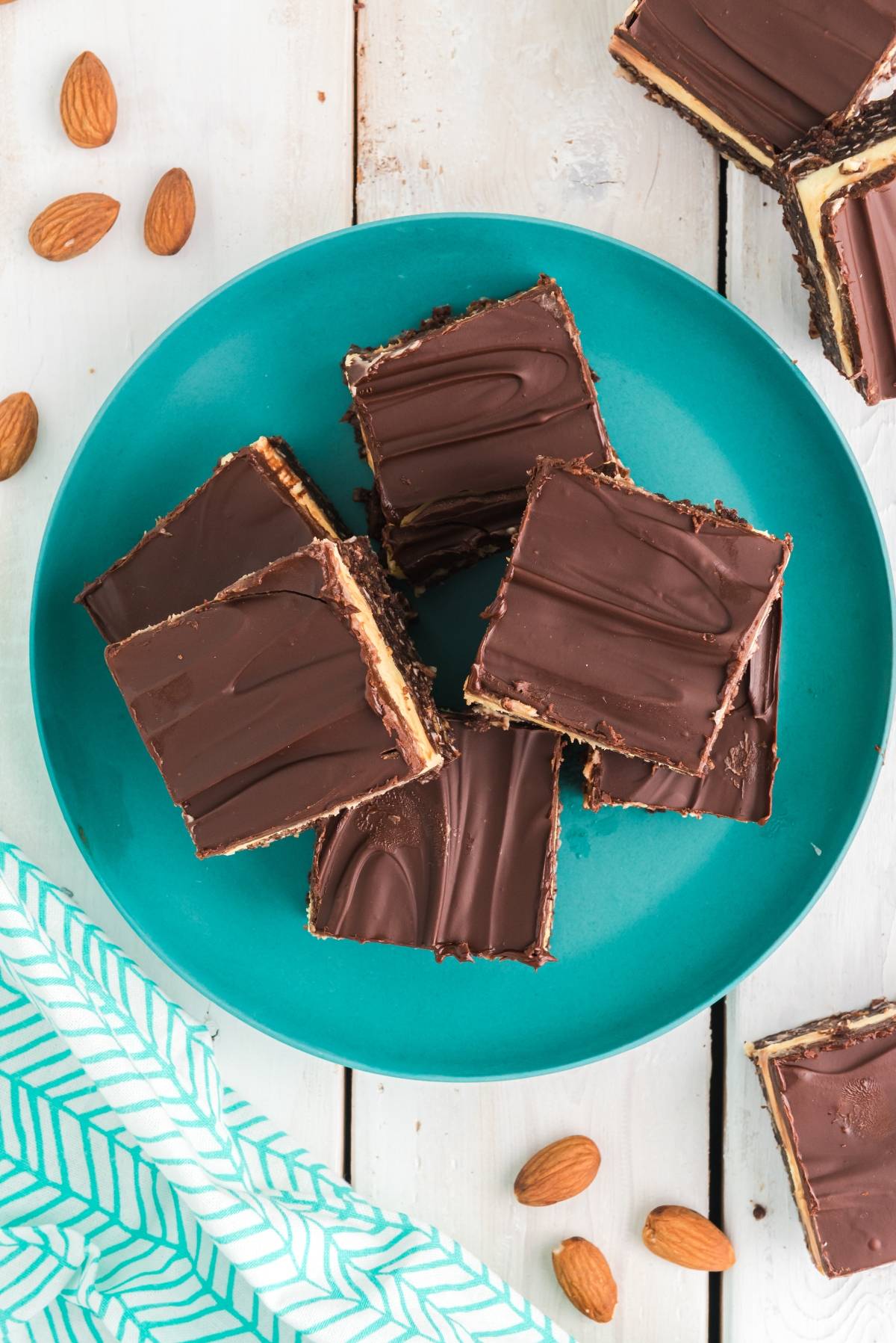 Six chocolate-covered dessert bars, inspired by a classic layered bar recipe, rest on a teal plate surrounded by whole almonds atop a white wooden table.