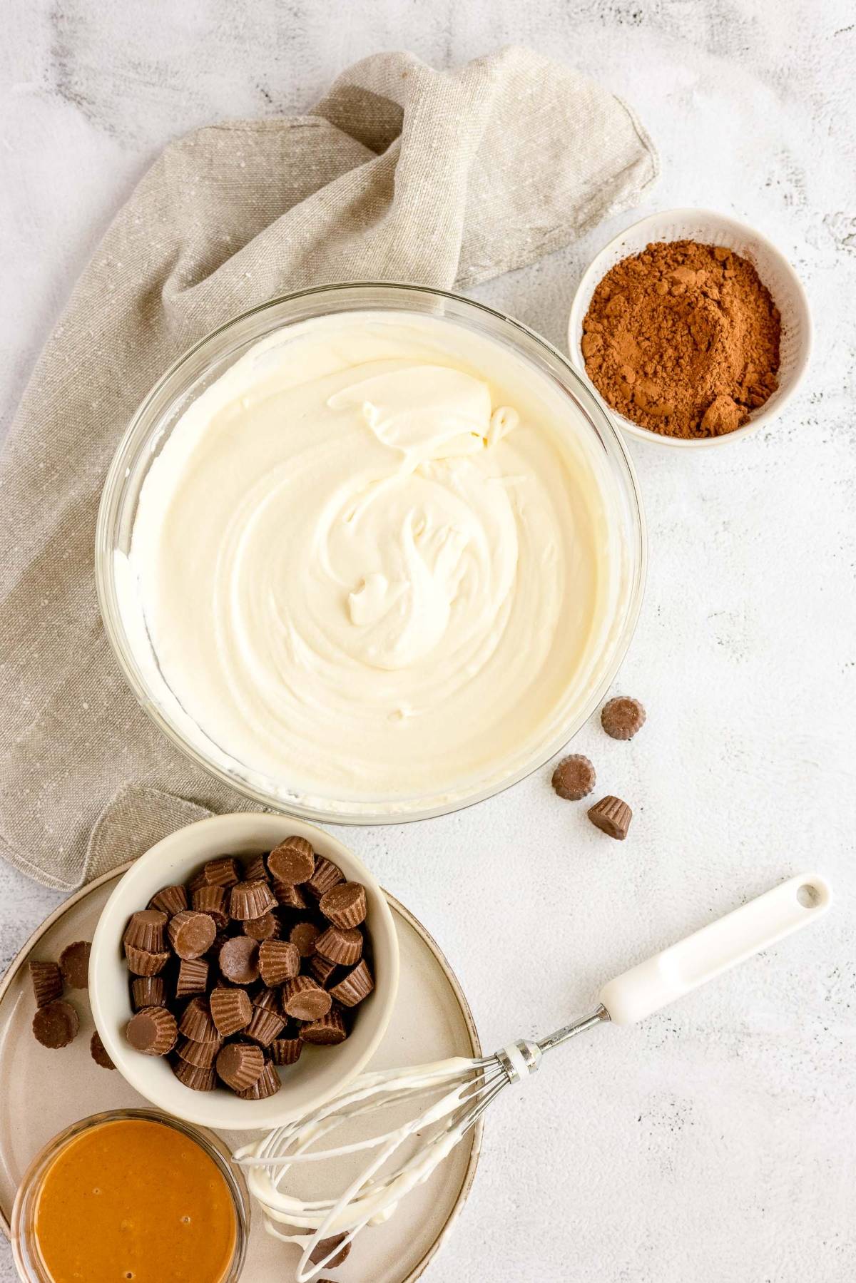 A bowl of creamy mixture, cocoa powder, mini peanut butter cups, caramel sauce, and a whisk on a table—perfect for making no churn peanut butter ice cream.
