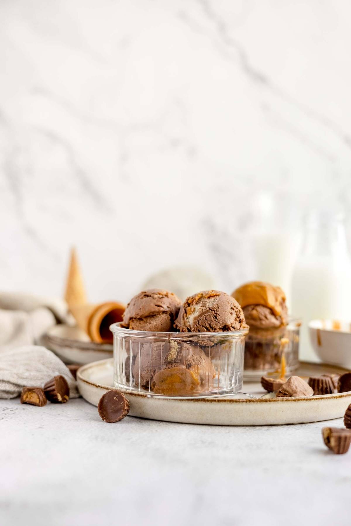 Two scoops of no churn chocolate peanut butter cup ice cream in a glass bowl, with peanut butter cups and an ice cream cone nearby.
