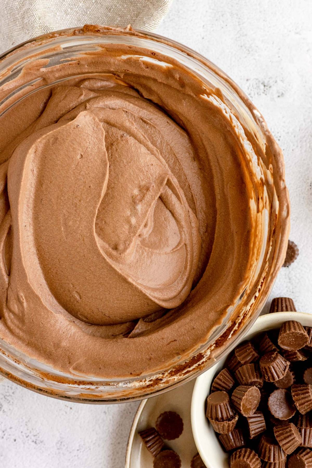 Bowl of creamy chocolate mousse next to a dish filled with mini peanut butter cups, perfect alongside no churn peanut butter ice cream on a light surface.