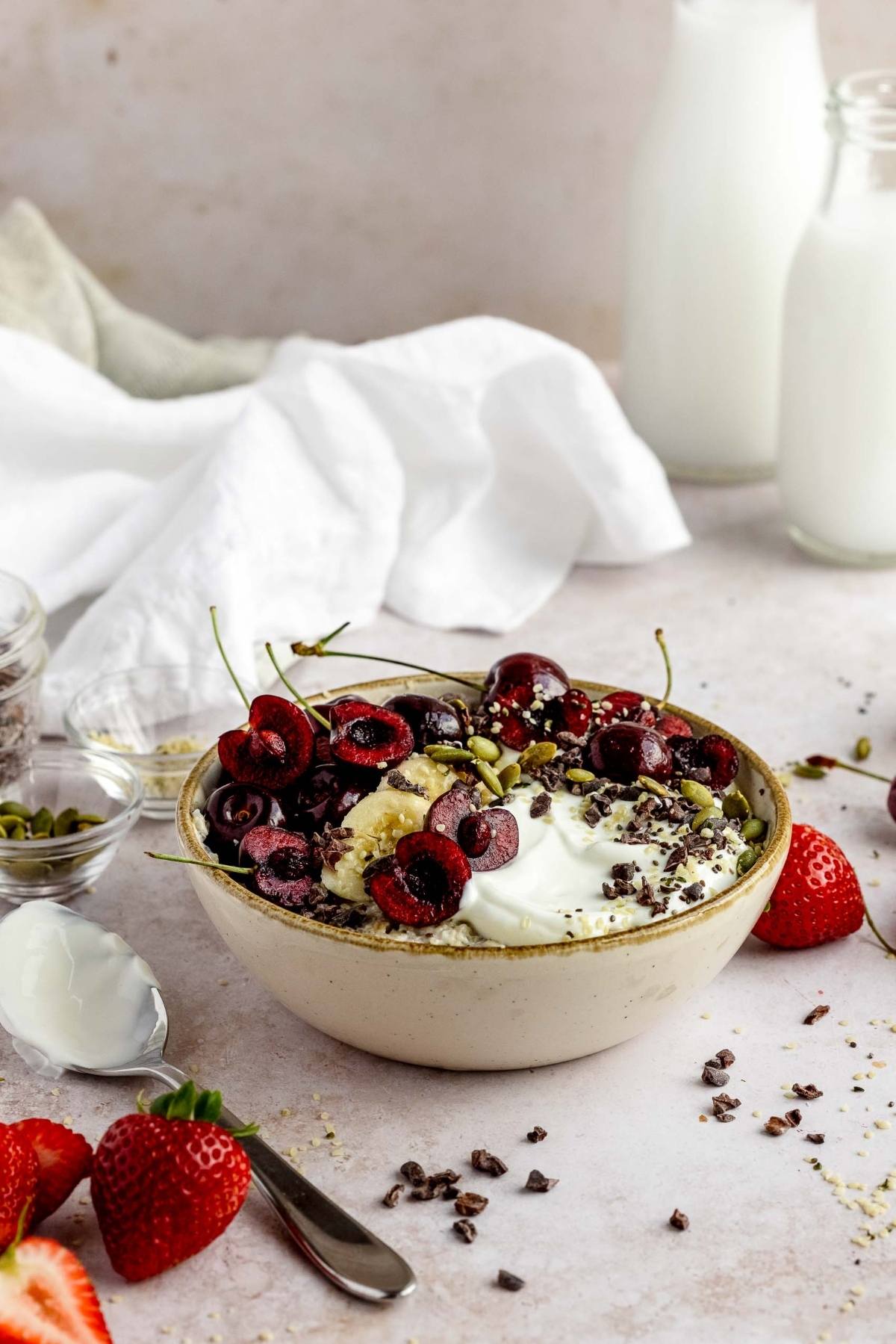 A bowl of yogurt topped with cherries, chocolate, and seeds, alongside milk bottles and strawberries—perfect inspiration for an oatmeal breakfast bowl or learning how to make an oatmeal bowl with fruit for breakfast.