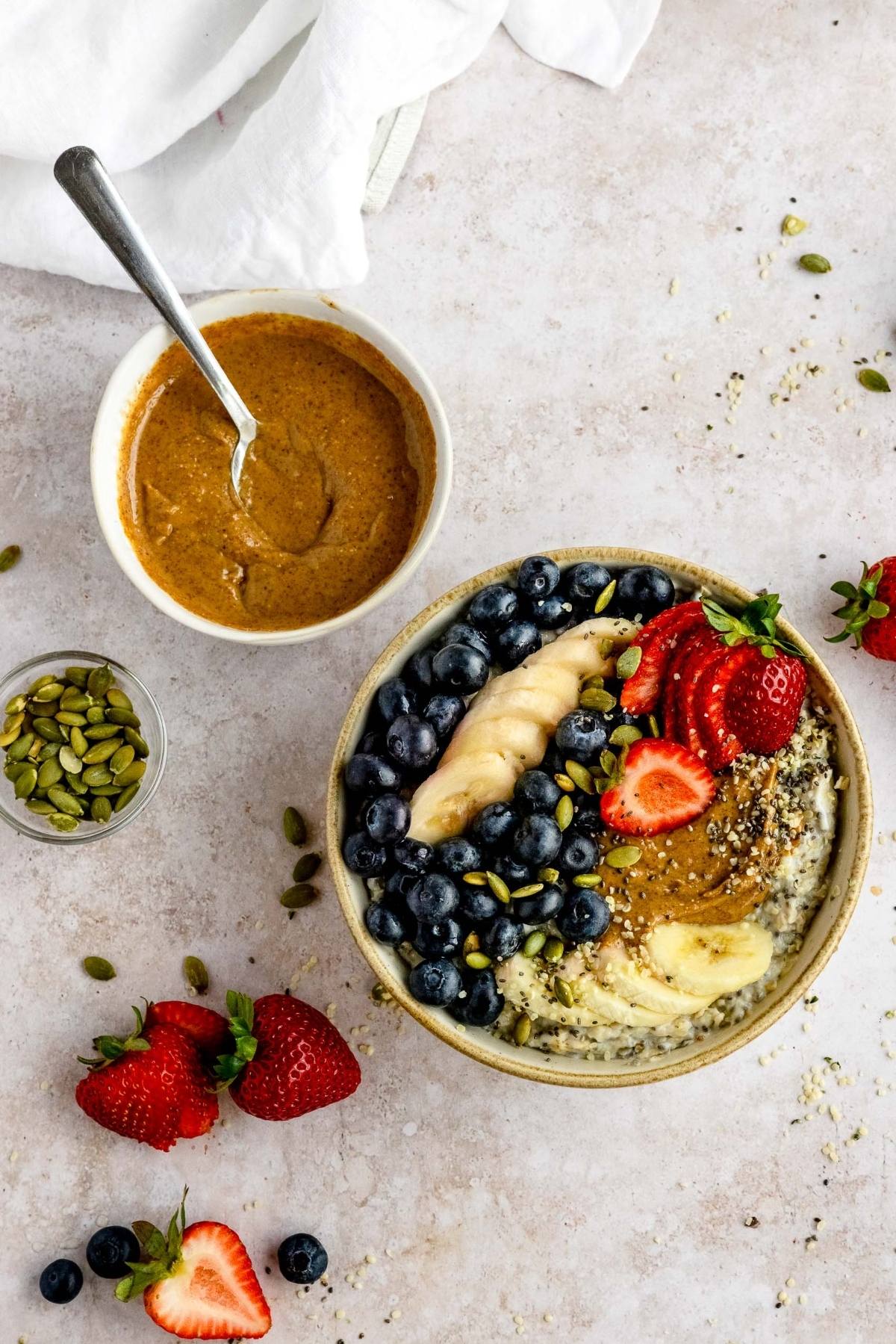 A healthy oatmeal breakfast bowl topped with blueberries, strawberries, banana, seeds, and a swirl of nut butter, with fresh ingredients nearby—a perfect example of delicious oatmeal bowl toppings.