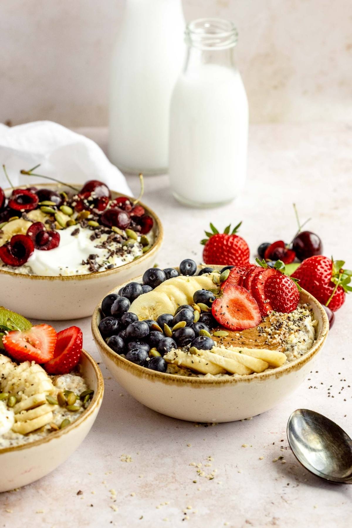 Three bowls of healthy oatmeal breakfast topped with fresh fruit, berries, and seeds sit beside bottles of milk and a spoon.