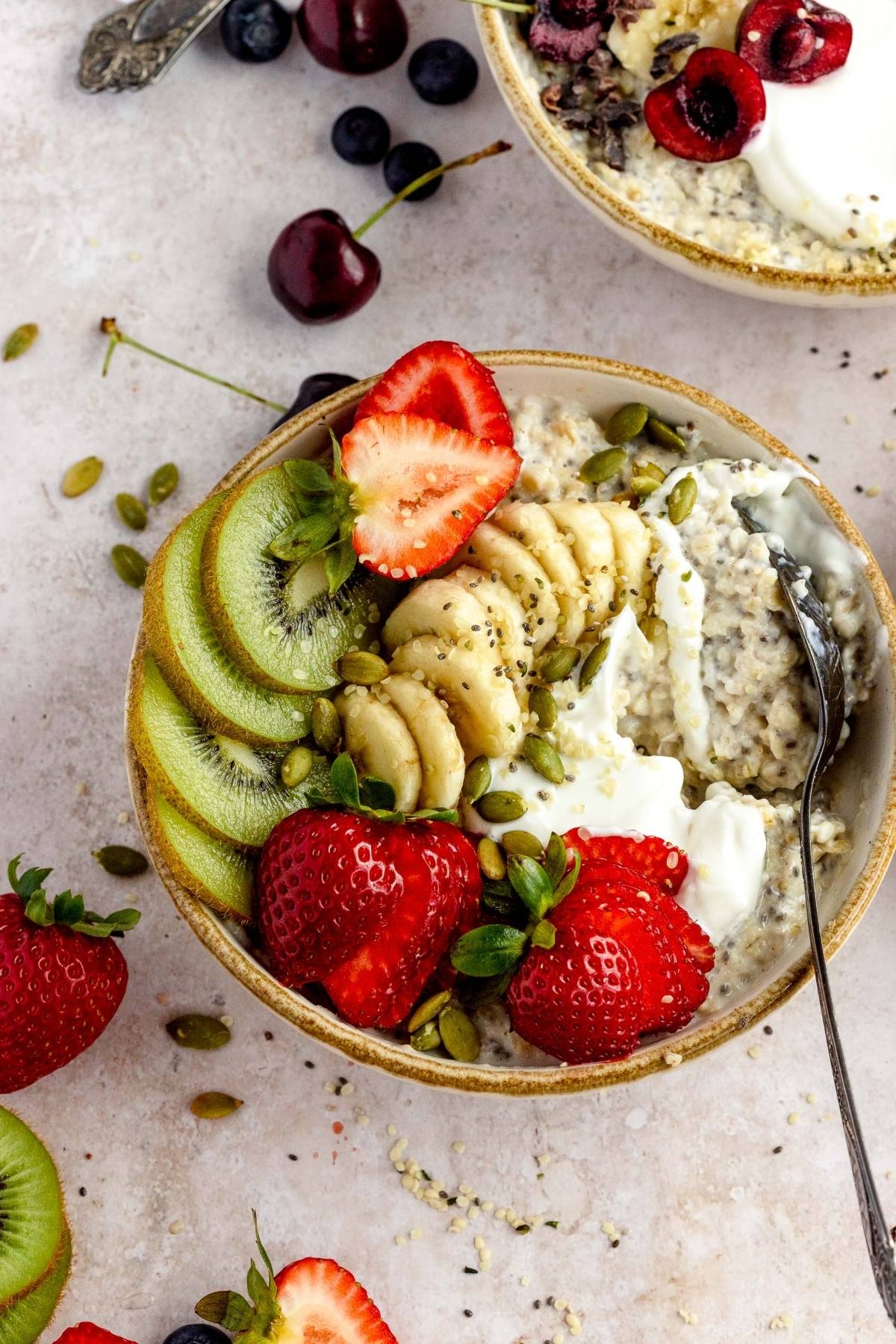 A bowl of oatmeal with banana, kiwi, strawberries, seeds, and yogurt makes a healthy oatmeal breakfast—perfect for anyone looking for oatmeal with fruit for breakfast. A spoon inside invites you to dig in and enjoy!.