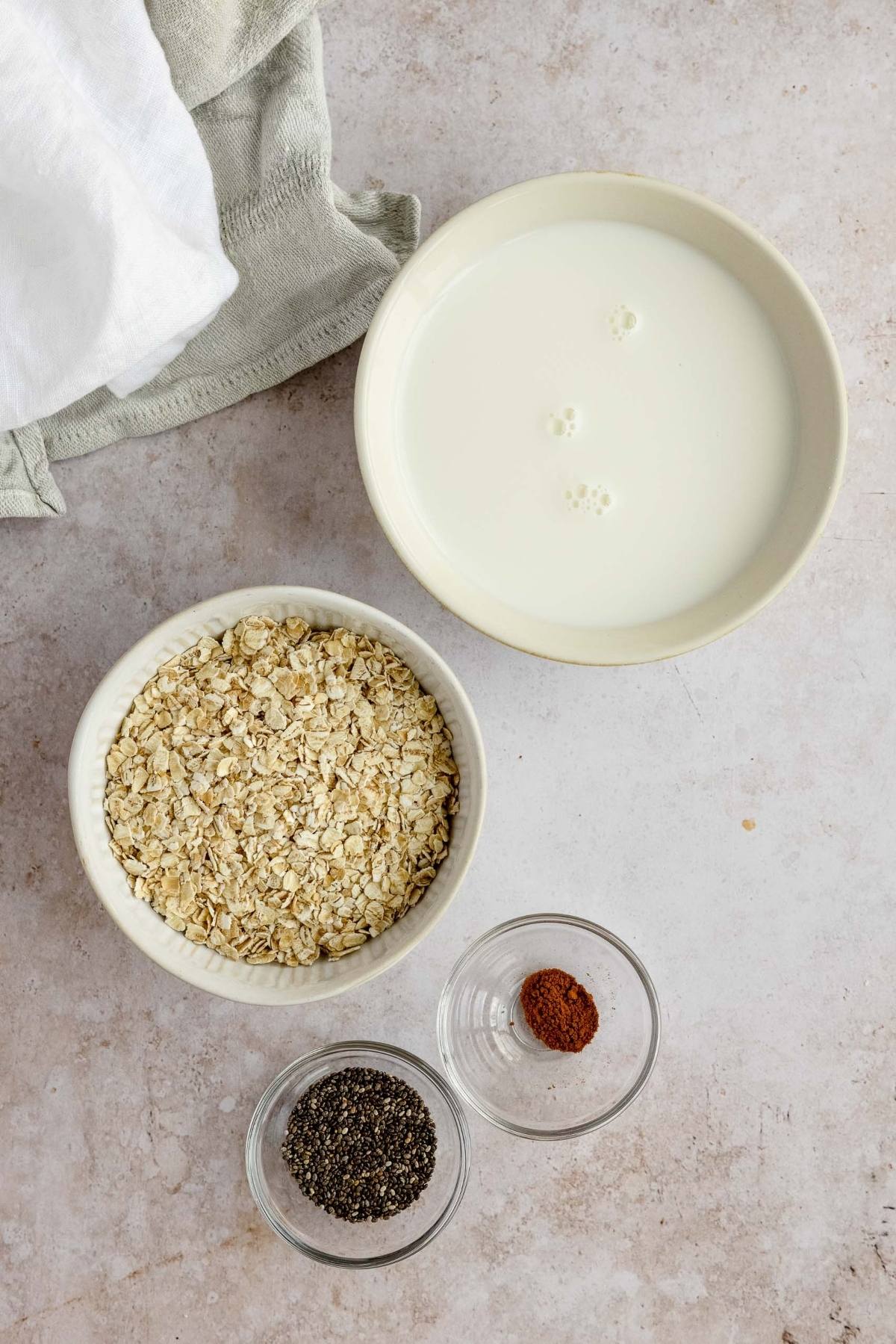 Bowls with milk, oats, chia seeds, and cinnamon on a light surface—perfect for an easy Oatmeal Breakfast Bowl. Towels rest in the corner.
