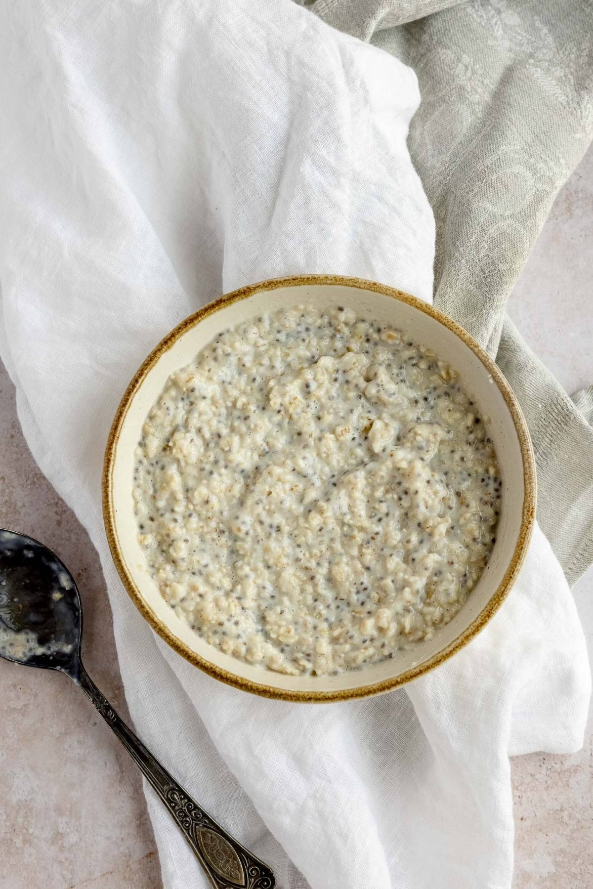 A delicious Oatmeal Breakfast Bowl with chia seeds sits on a white cloth, next to a spoon—perfect for adding your favorite oatmeal bowl toppings.