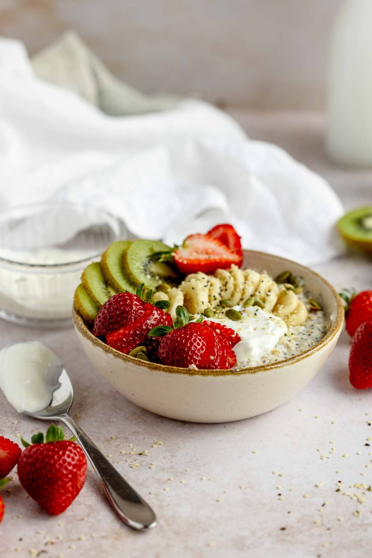 A bowl of healthy oatmeal breakfast topped with sliced banana, kiwi, strawberries, yogurt, and chia seeds sits on a table.