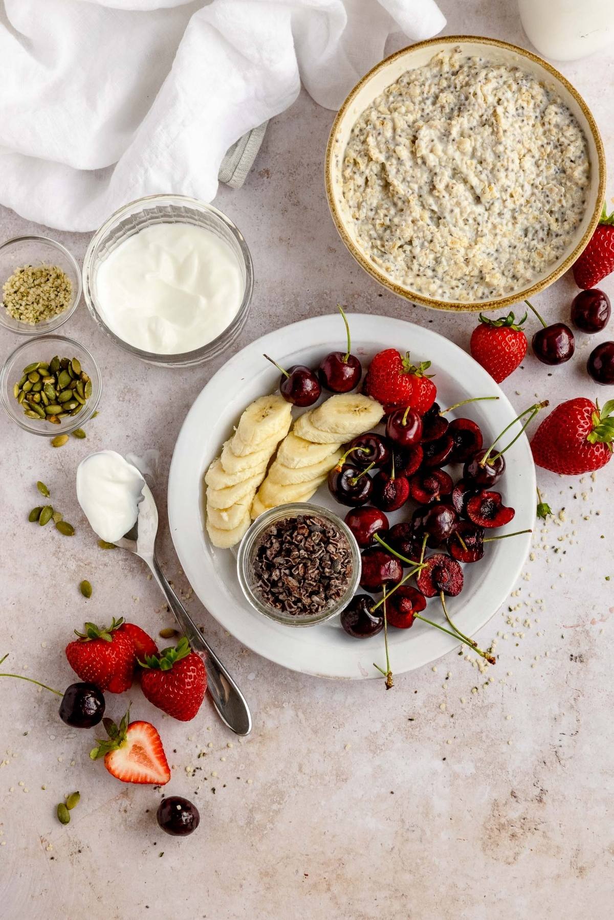 A plate with sliced banana, cherries, cacao nibs, strawberries, yogurt, and a bowl of healthy oatmeal breakfast nearby.