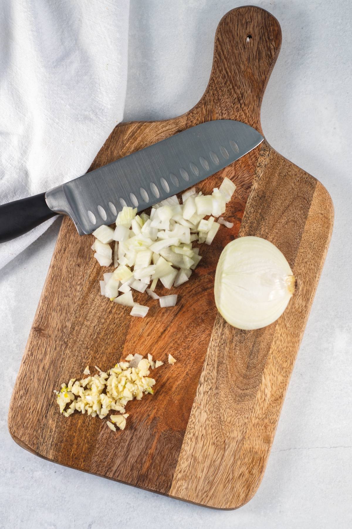 Chopped onion, minced garlic, and a knife on a wooden cutting board with a white towel nearby—perfect prep for an easy spinach pasta or fresh greens vegan pasta dinner.