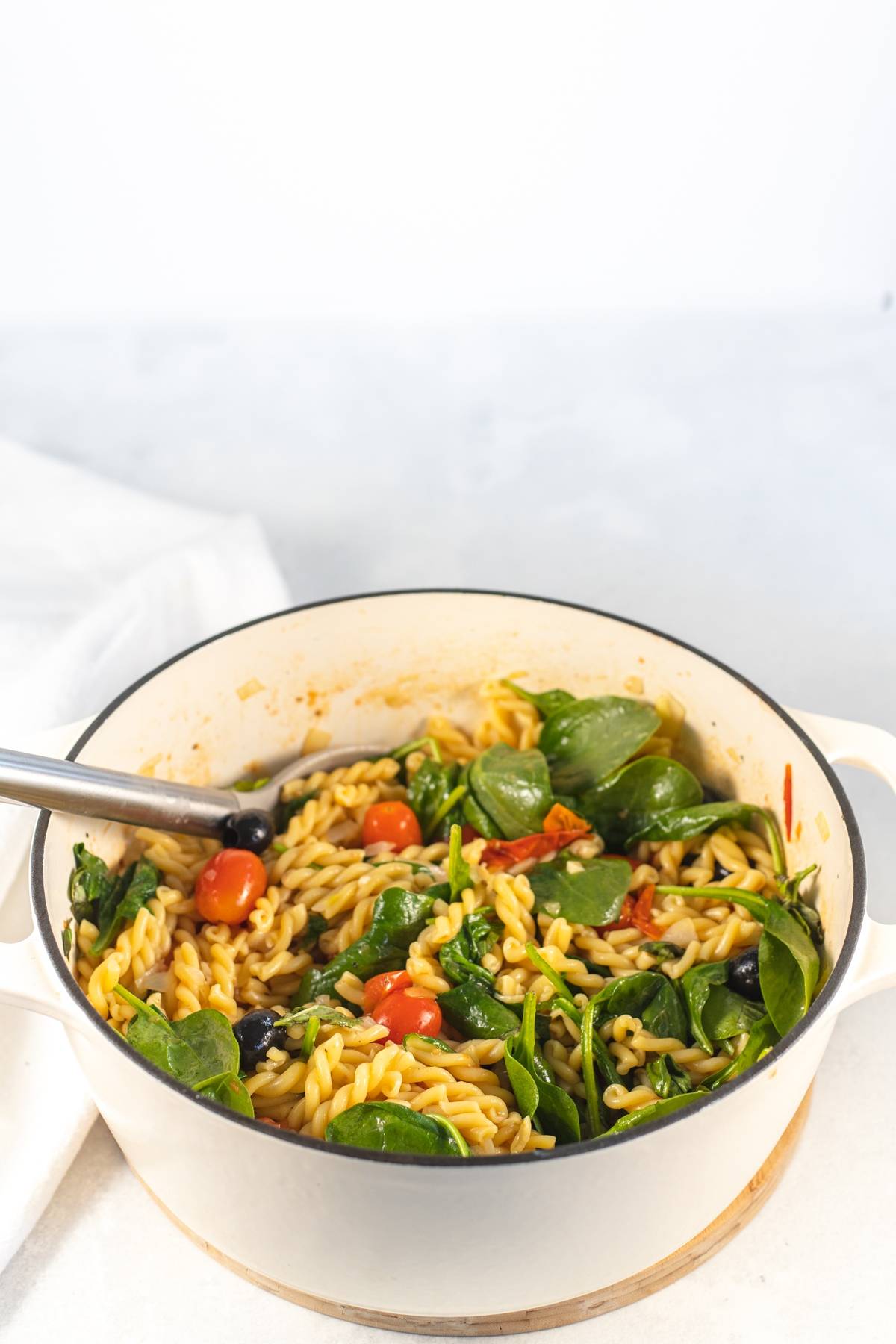 One Pot Pasta with baby spinach and cherry tomatoes, tossed with olives and served in a large pot on a white surface.