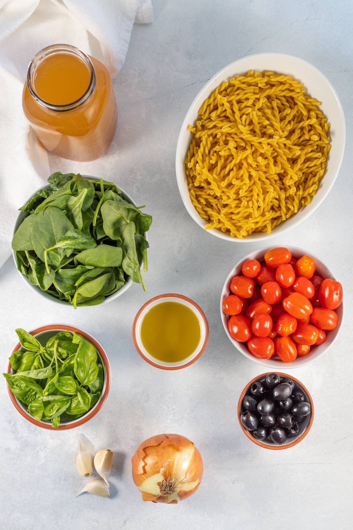 Top-down view of easy spinach pasta ingredients—pasta with spinach and cherry tomatoes, olives, olive oil, broth, basil, garlic, and onion—all laid out on a light surface.