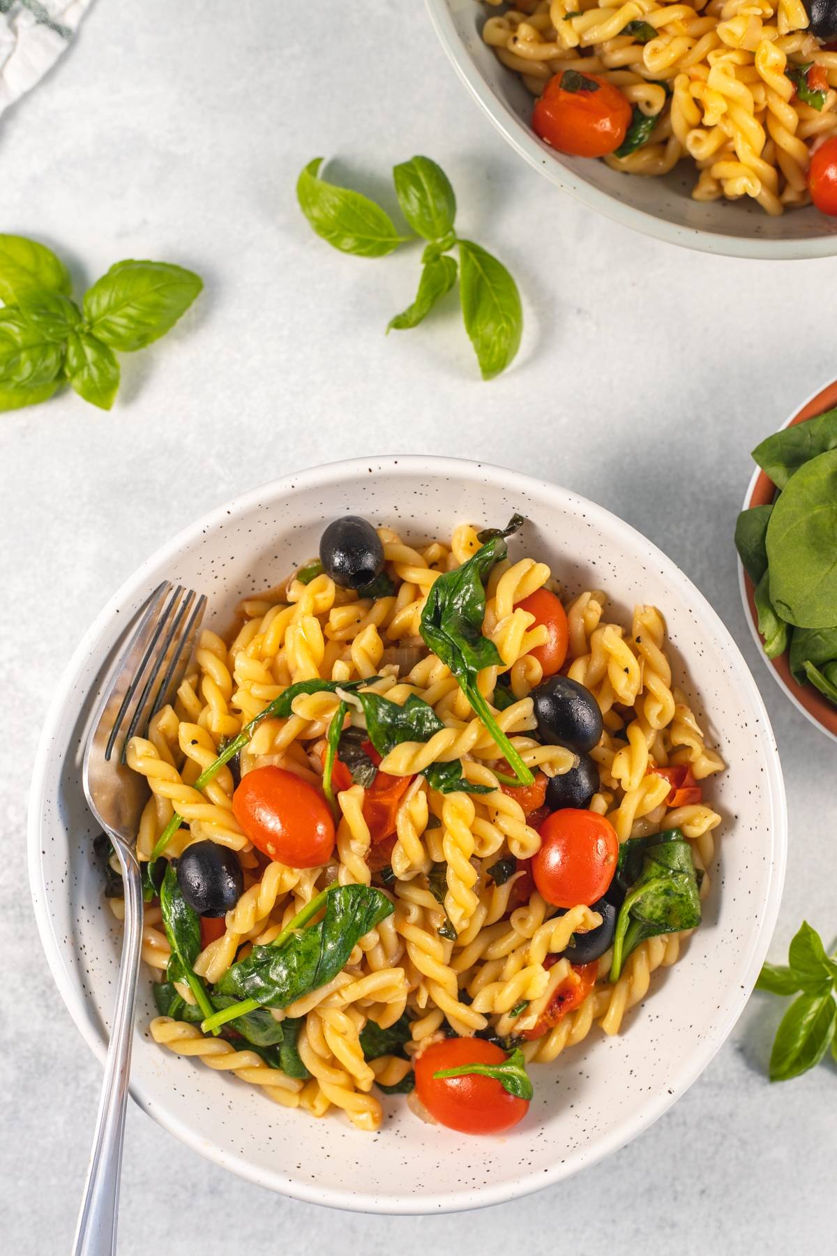 A bowl of easy spinach pasta with cherry tomatoes, olives, and a fork on a light surface, garnished with fresh basil leaves nearby.