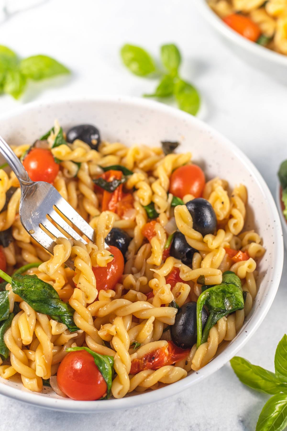 A bowl of easy spinach pasta with cherry tomatoes, black olives, and a fork, garnished with fresh basil—perfect for a vibrant meal.