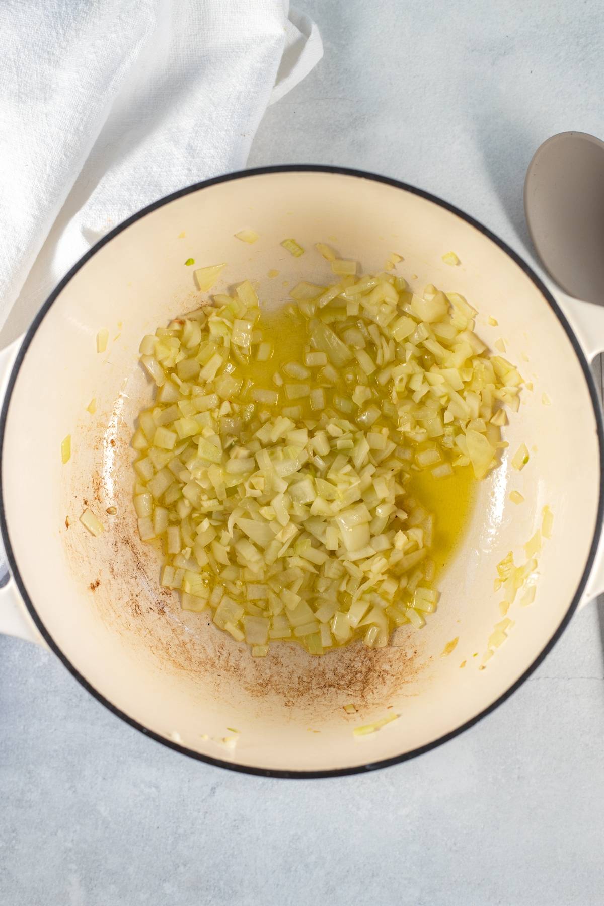 Chopped onions sautéing in a white pot with oil, next to a spoon and a white towel, set the stage for an easy spinach pasta—just add baby spinach and cherry tomatoes for a tasty One Pot Pasta on a light surface.