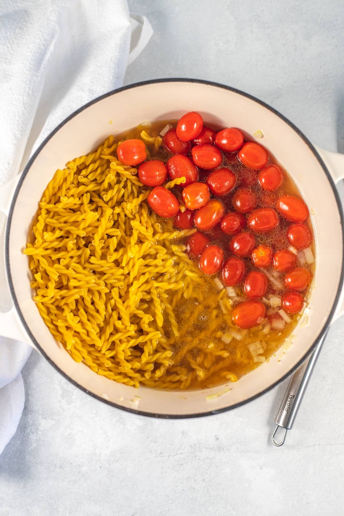 A pot with uncooked One Pot Pasta, baby spinach and cherry tomatoes, and broth, ready to be cooked on a light countertop.