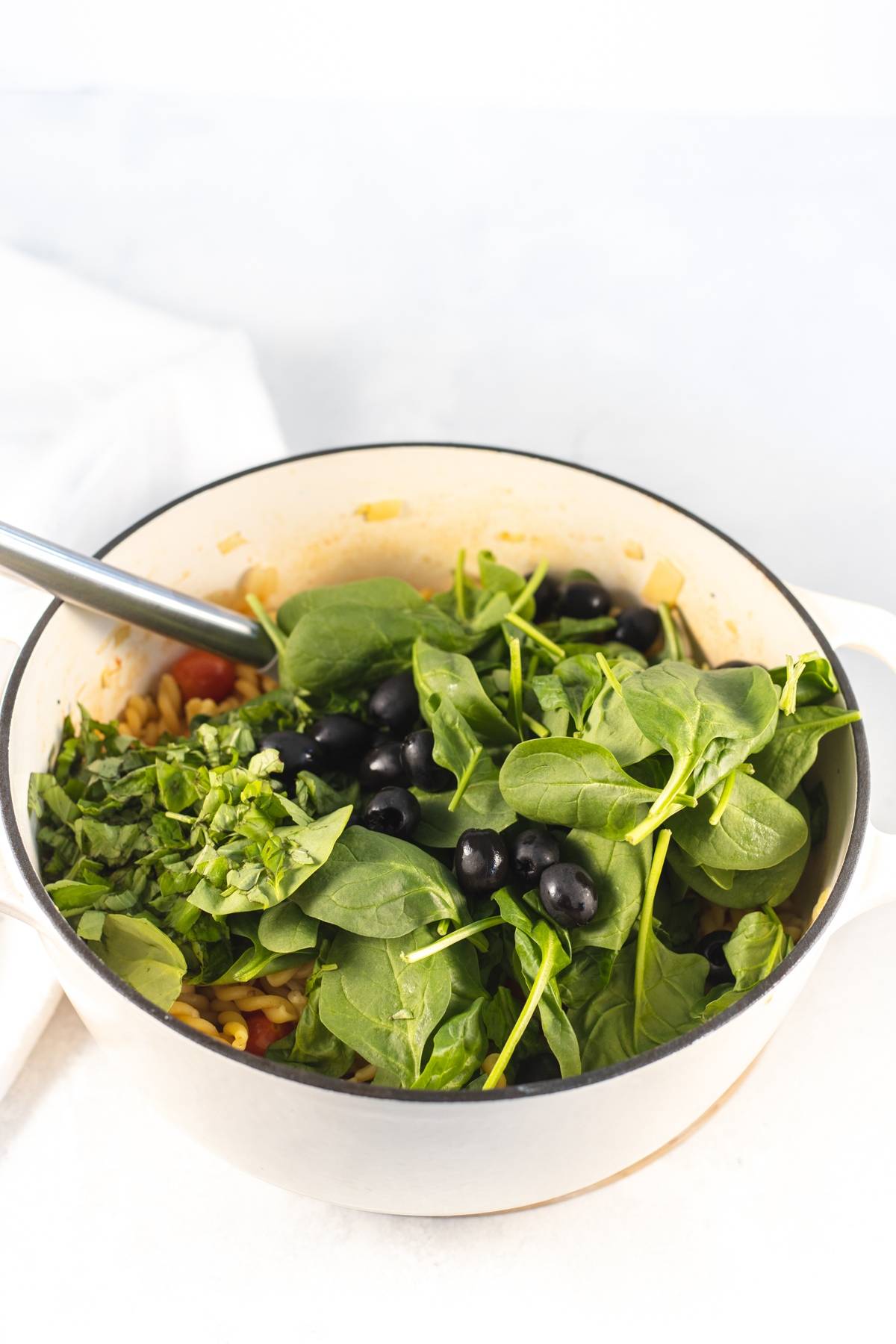 A white pot filled with fresh baby spinach, cherry tomatoes, black olives, and pasta, all brought together as a vibrant One Pot Pasta, with a metal spoon inside.