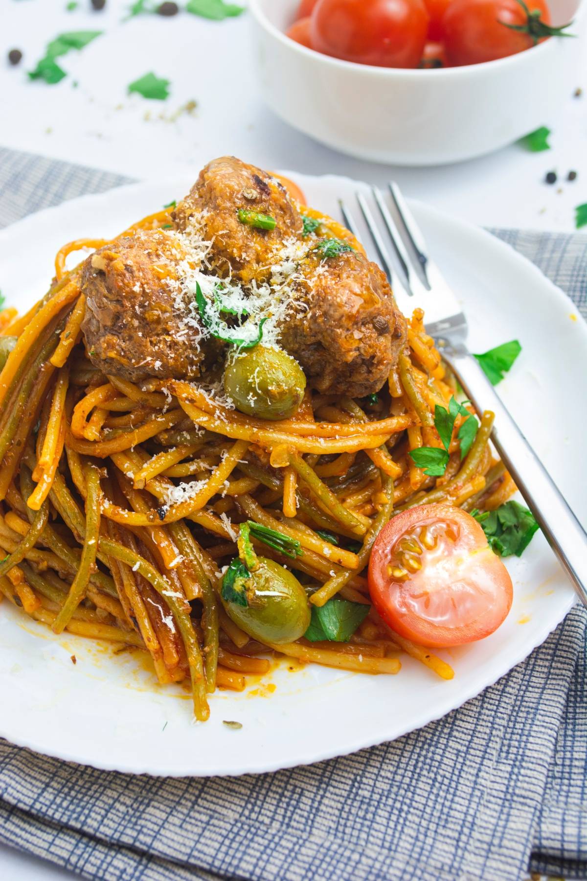 Mediterranean spaghetti and meatballs with green olives and grated cheese, garnished with parsley and sliced tomato, served on a white plate. An easy one pot pasta perfect for weeknight dinners.