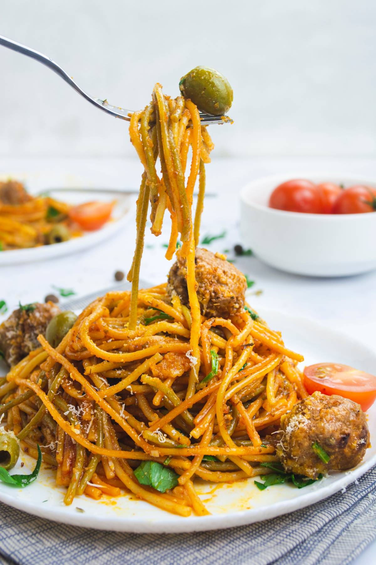A fork twirls easy one pot pasta with spaghetti, meatballs, and an olive on a plate, garnished with herbs and a tomato wedge.