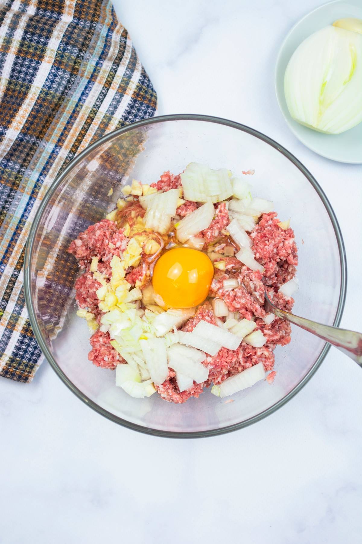 A glass bowl with ground meat, chopped onions, and an egg yolk—perfect for preparing one pot Mediterranean pasta—sits beside a striped towel and a plate with onion.
