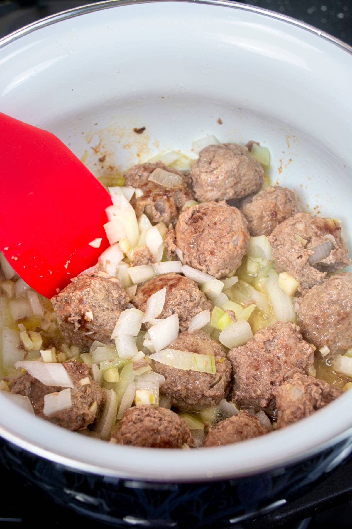 Meatballs and chopped onions being stirred with a red spatula in a white pot, perfect for making an easy one pot pasta or Mediterranean spaghetti and meatballs.