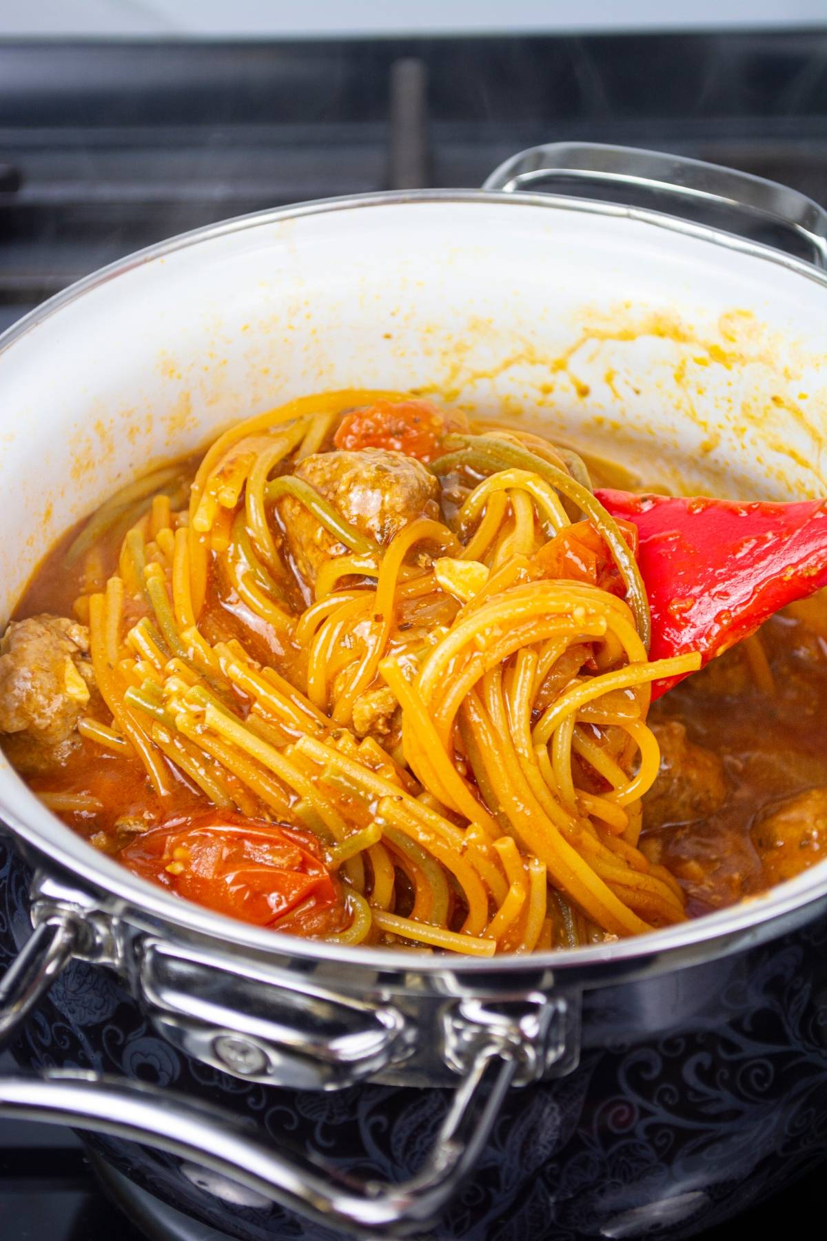 Easy one pot pasta with meatballs and tomatoes simmers in a pot, stirred gently with a red spatula.