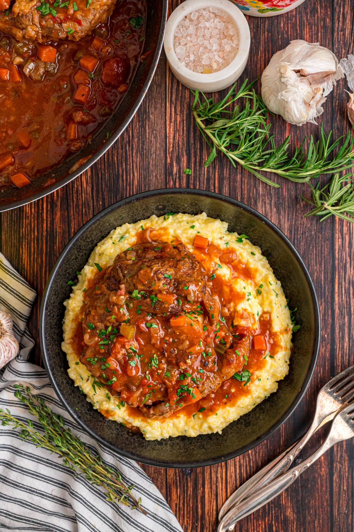 A bowl of polenta topped with braised osso buco and vegetables, surrounded by herbs and seasonings on a wooden table.