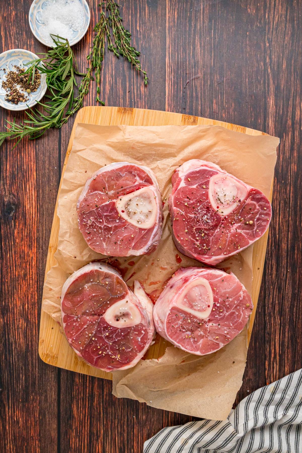 Four raw osso buco beef cuts with bone marrow on parchment paper, seasoned with pepper, on a wooden board.