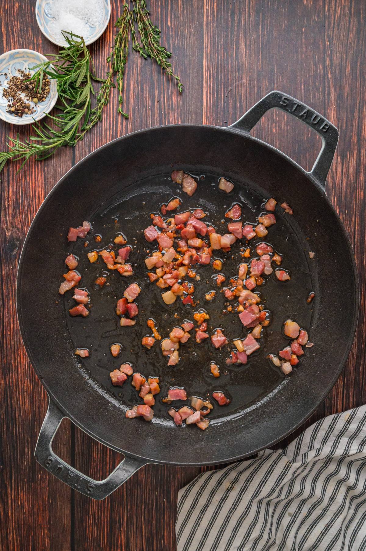 Diced bacon cooking in a black skillet on a wooden surface with herbs and seasoning nearby.