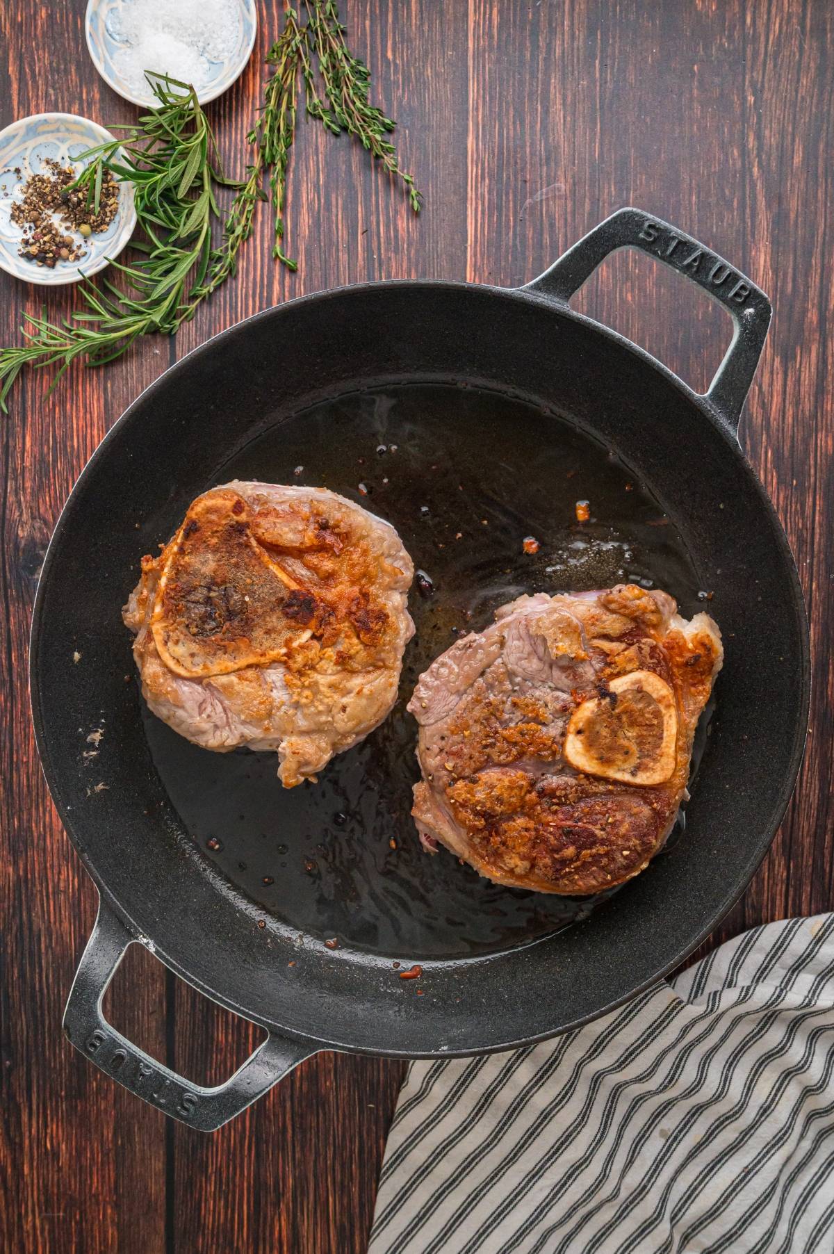 Two bone-in steaks searing in a black skillet on a wooden surface, with herbs and seasonings nearby.