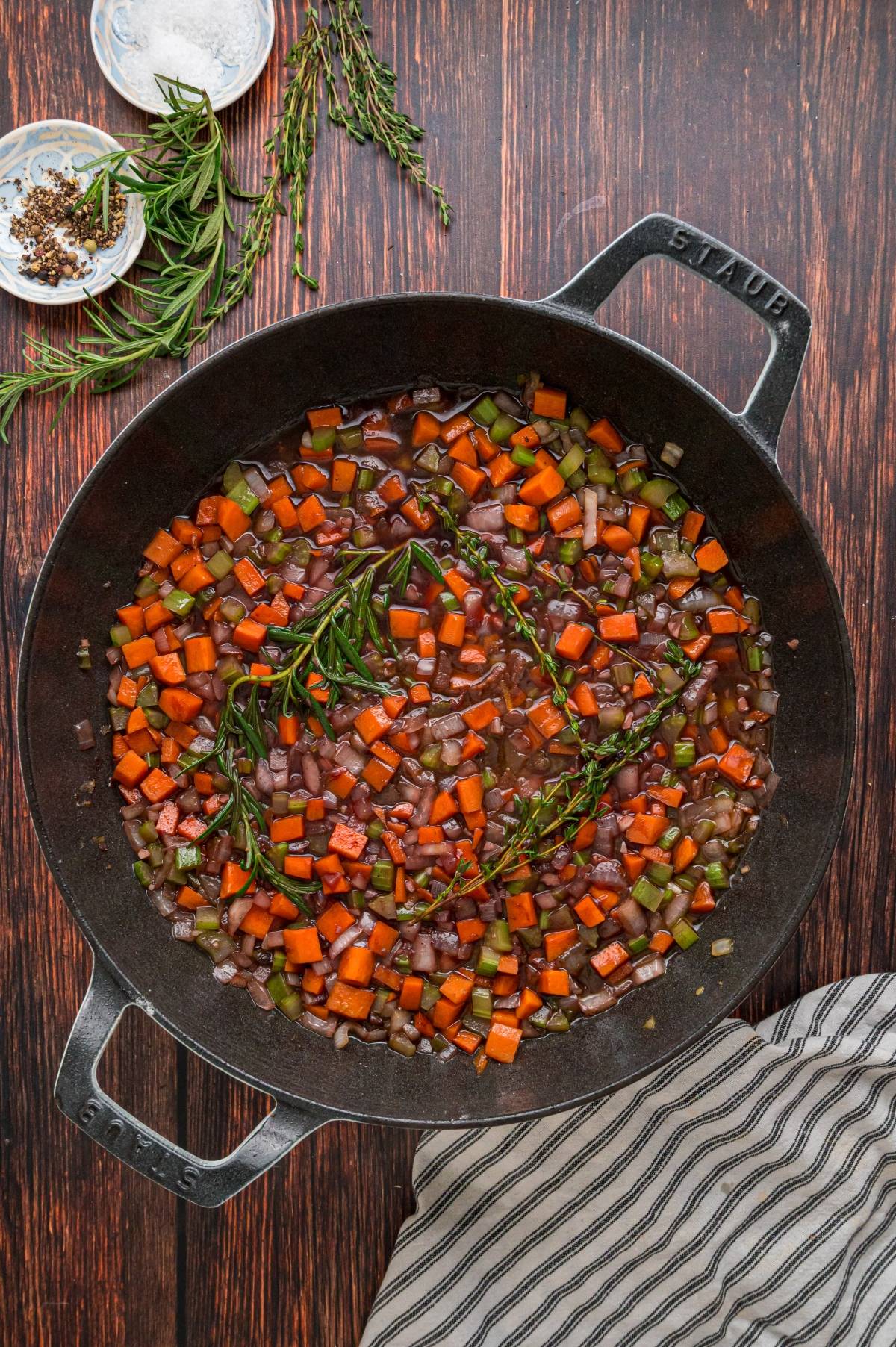 Top view of a skillet with chopped carrots, onions, celery, and herbs simmering in a dark sauce on a wooden table.