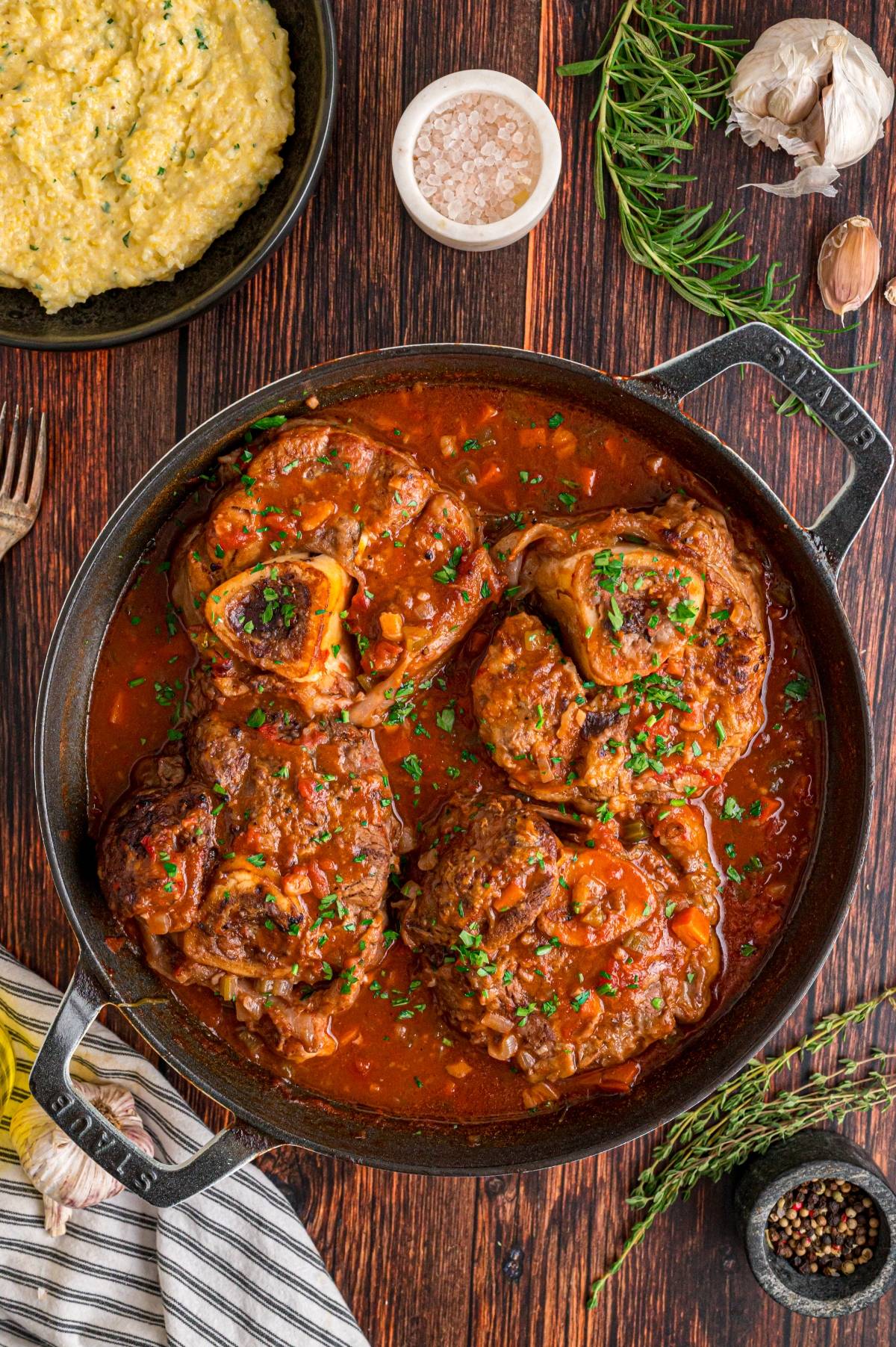 A pan of braised osso buco in tomato sauce, garnished with herbs, beside mashed polenta and seasonings.