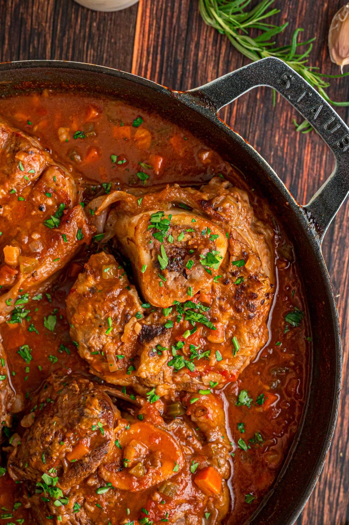 Braised beef in tomato sauce topped with chopped herbs in a black pot on a wooden table.