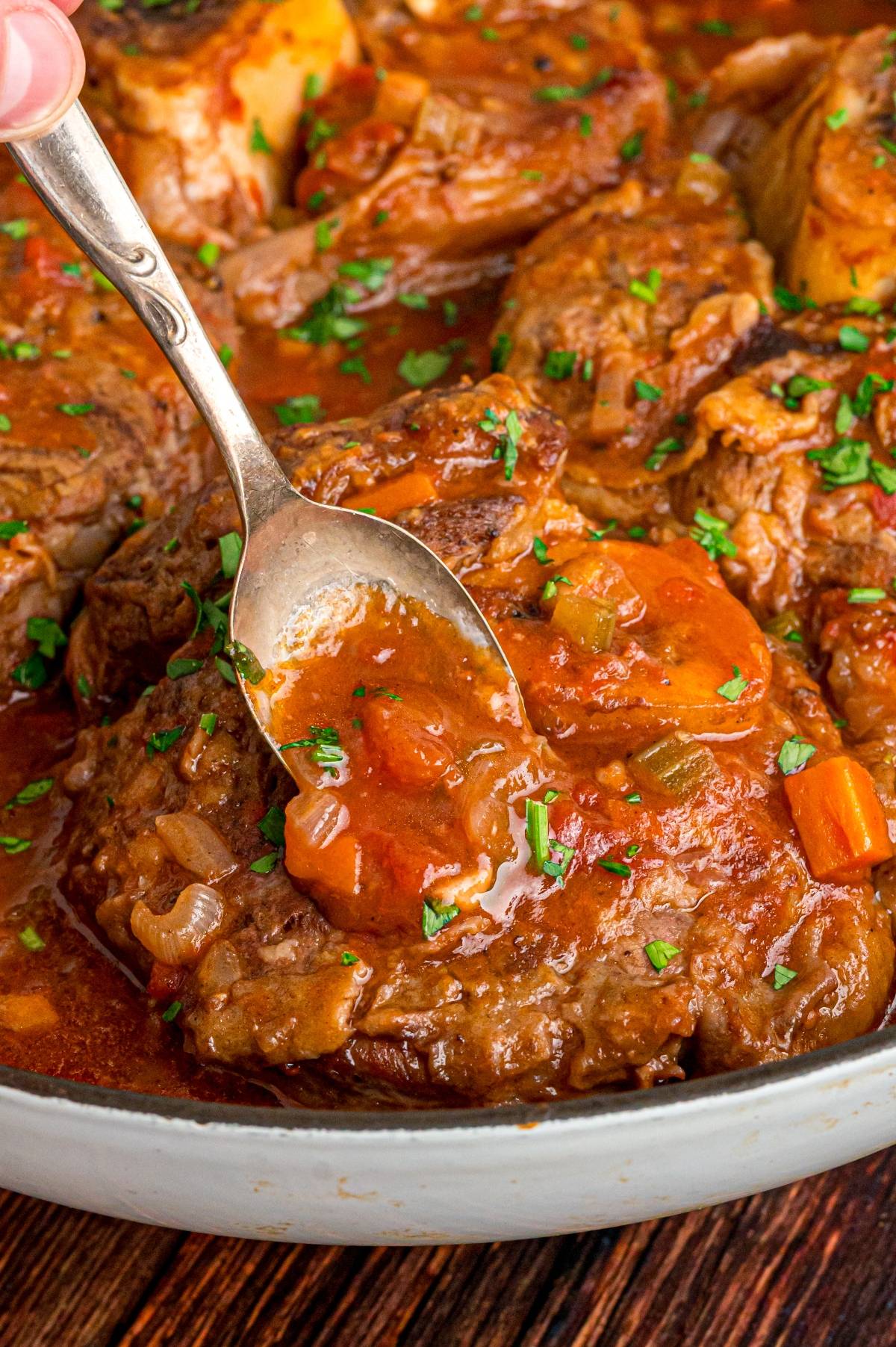 A spoon scoops sauce over braised meat with carrots, celery, and herbs in a rich, red stew.