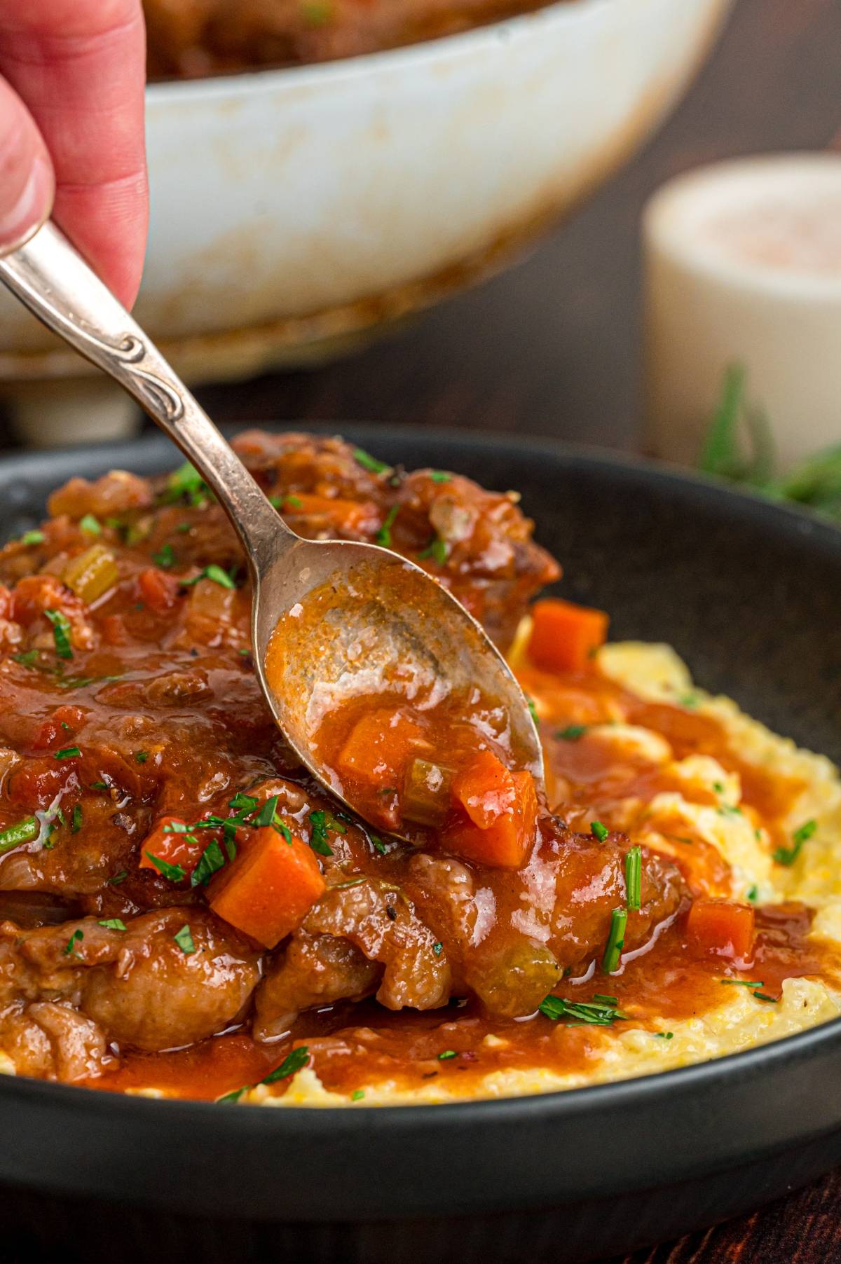 A spoon serving veal shank with carrots and sauce over creamy polenta in a black bowl.