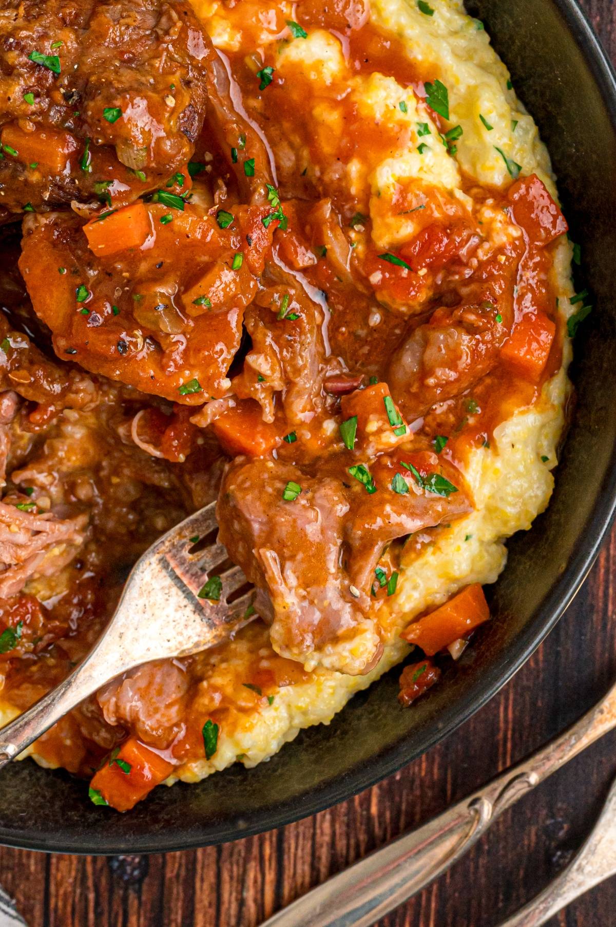 Italian osso buco with carrots and sauce served over creamy mashed potatoes, with a fork picking up a bite.