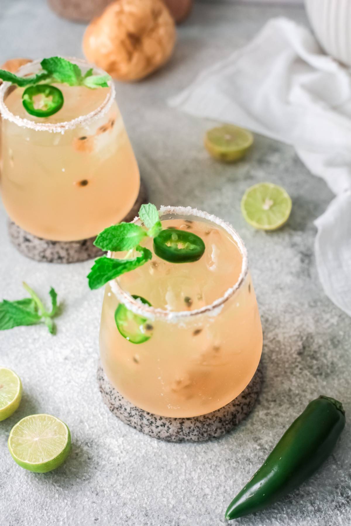 Two cocktails with lime, mint, jalapeño slices, and salted rims on a gray surface with fresh limes and jalapeño—perfect for trying an easy margarita recipe or adding a twist with passion fruit and lime flavors.