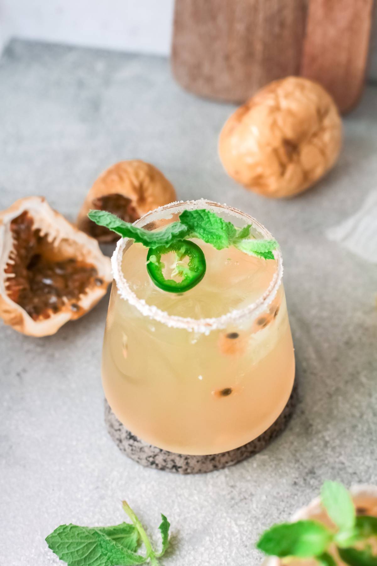 A passion fruit cocktail garnished with mint and jalapeño sits on a coaster, with fresh passion fruits and mint nearby—an easy margarita recipe perfect for any occasion.