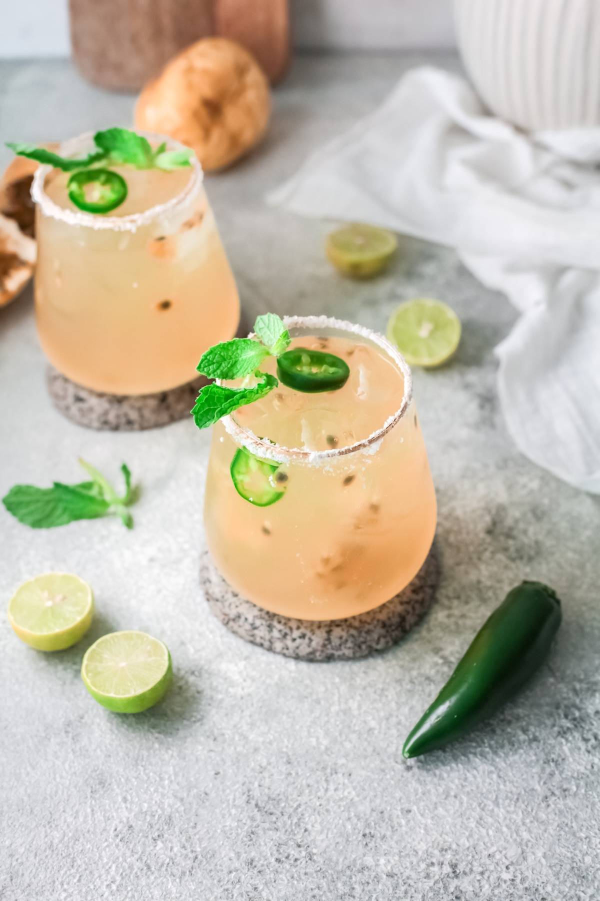 Two glasses of a light cocktail with ice, garnished with mint and jalapeño slices, next to lime halves—an easy margarita recipe that's perfect for a fresh fruit margarita twist.
