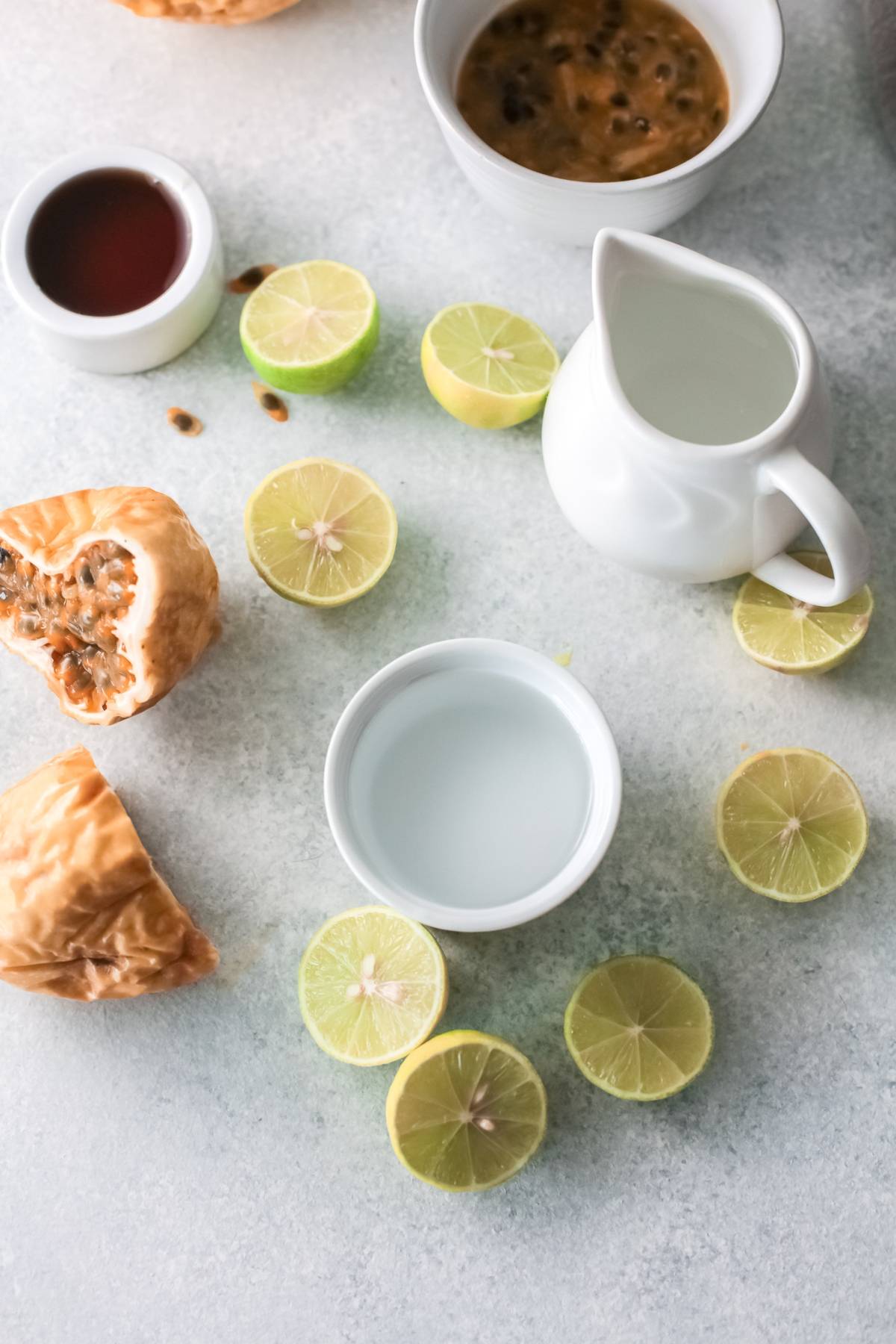 Sliced limes, a pitcher of liquid, bowls of passion fruit and lime, and syrup arranged on a light surface—perfect for an easy margarita recipe.