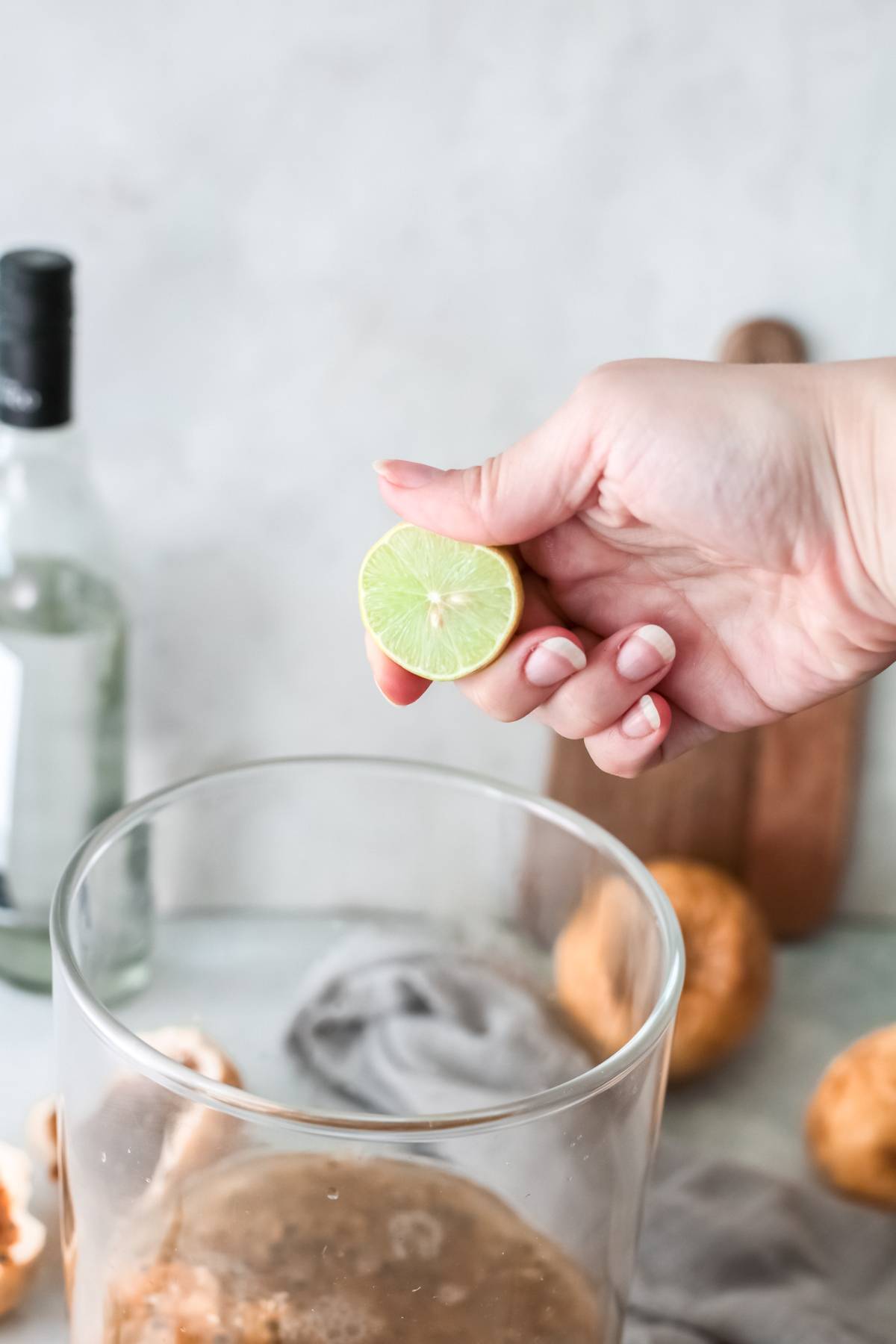 A hand holds half a lime above a glass, ready to squeeze it for an easy margarita recipe; ingredients for a passion fruit margarita are visible in the background.