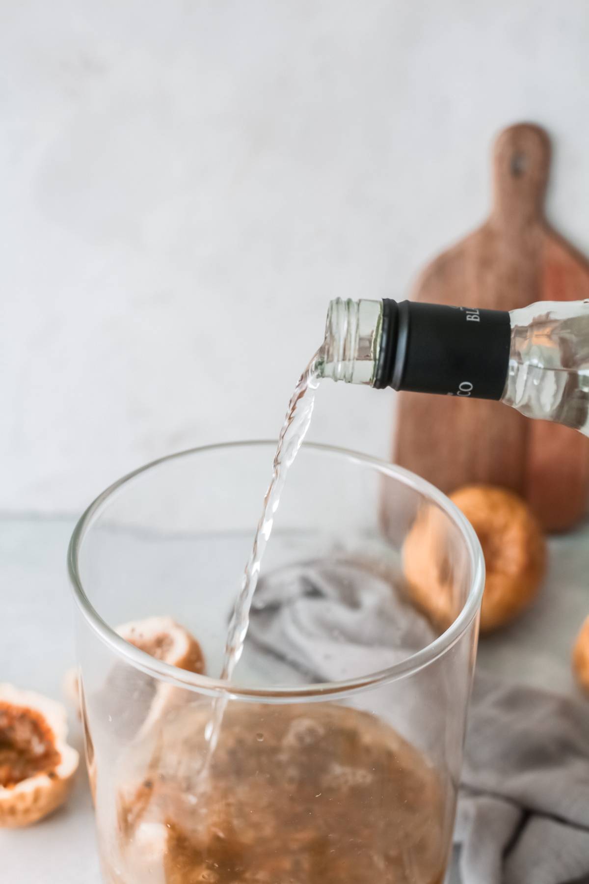 Clear liquid being poured from a bottle into a glass, with fresh fruit and a wooden board in the background—perfect inspiration for an easy margarita recipe.