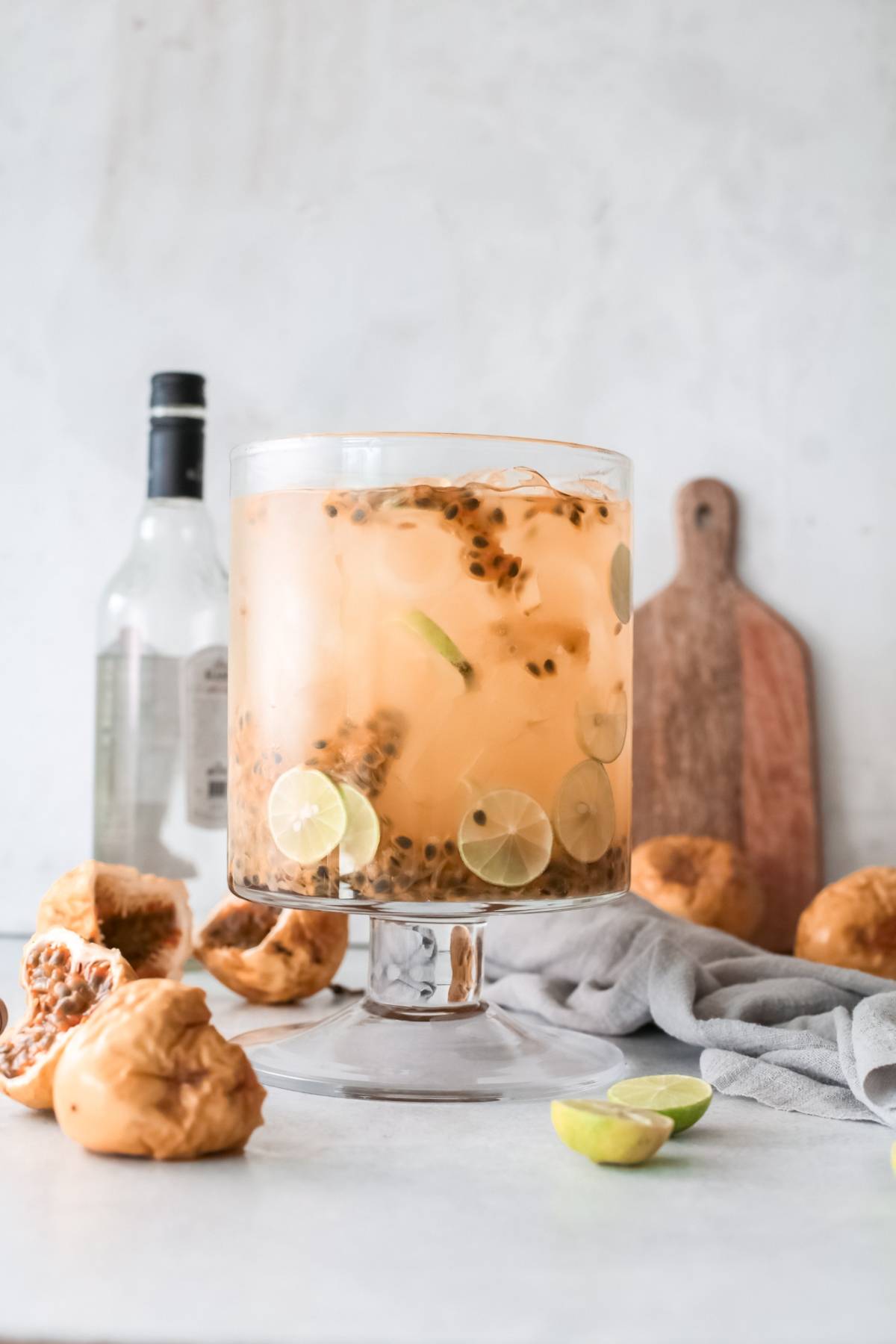 Large glass bowl of punch with lime slices and passionfruit, surrounded by limes, passionfruit, and a bottle—perfect for serving as a refreshing passion fruit margarita at your next gathering.