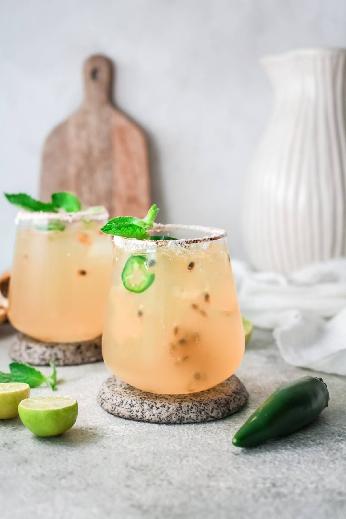 Two light orange passion fruit cocktails with ice, mint, and sliced jalapeño, garnished with a salt rim and served on stone coasters—perfect for lovers of a fresh fruit margarita.
