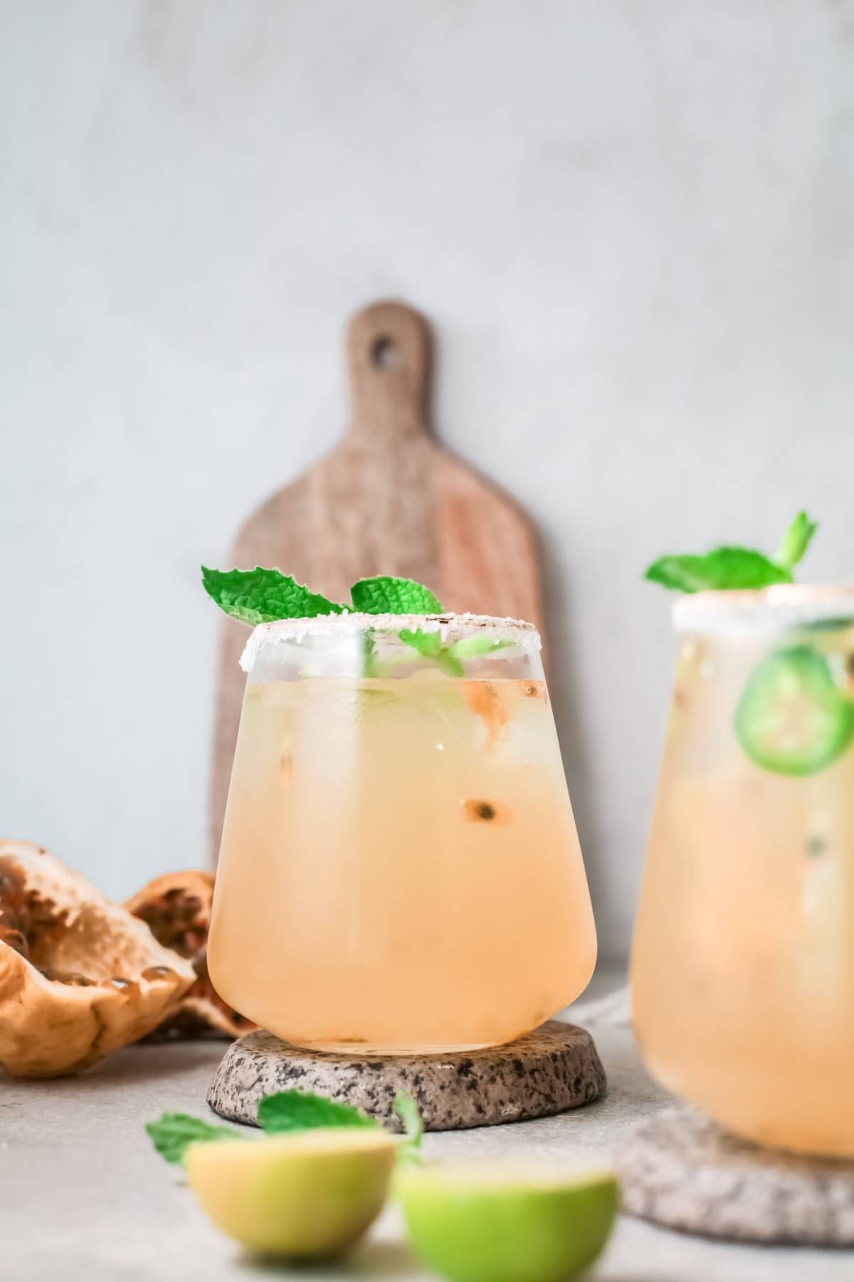 A light-colored passion fruit cocktail in a glass with mint garnish, salted rim, and limes, with a cutting board in the background.