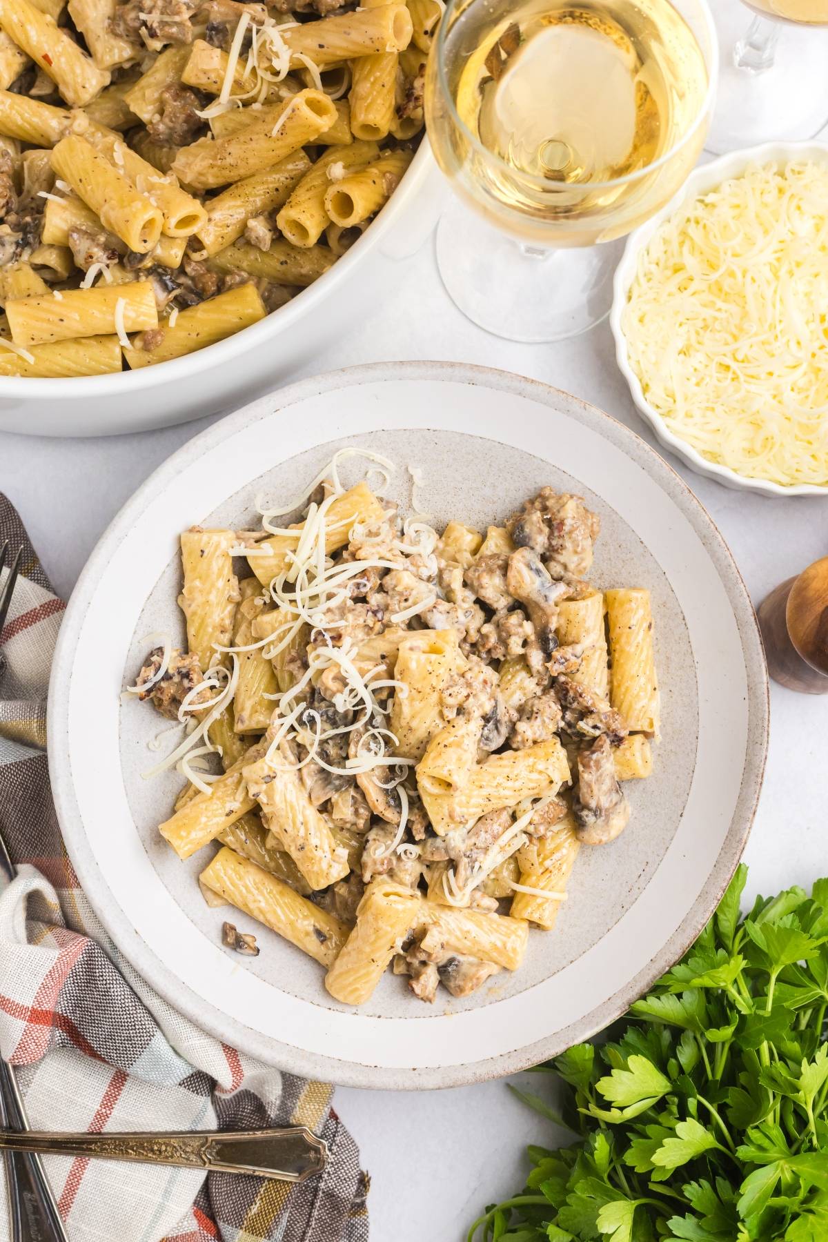 Plate of rigatoni pasta with sausage, mushrooms, and grated cheese, next to wine, parsley, and a bowl of extra pasta.