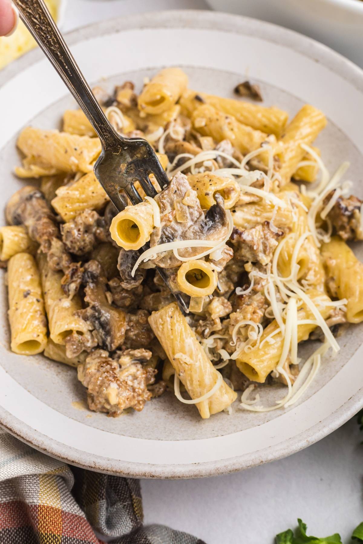 A fork lifts cheesy rigatoni pasta with mushrooms and ground meat from a plate.